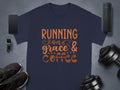 A navy blue Graphic T-Shirt by Garment Graphics features vibrant orange text, Running on Grace & Coffee. Its stylishly showcased with a smartphone, smartwatch, black sneakers, dumbbells, and a portable speaker on a sleek gray background.