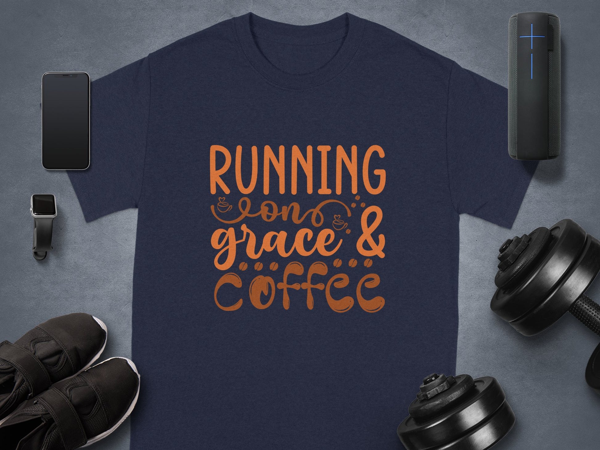 A navy blue Graphic T-Shirt by Garment Graphics features vibrant orange text, Running on Grace & Coffee. Its stylishly showcased with a smartphone, smartwatch, black sneakers, dumbbells, and a portable speaker on a sleek gray background.