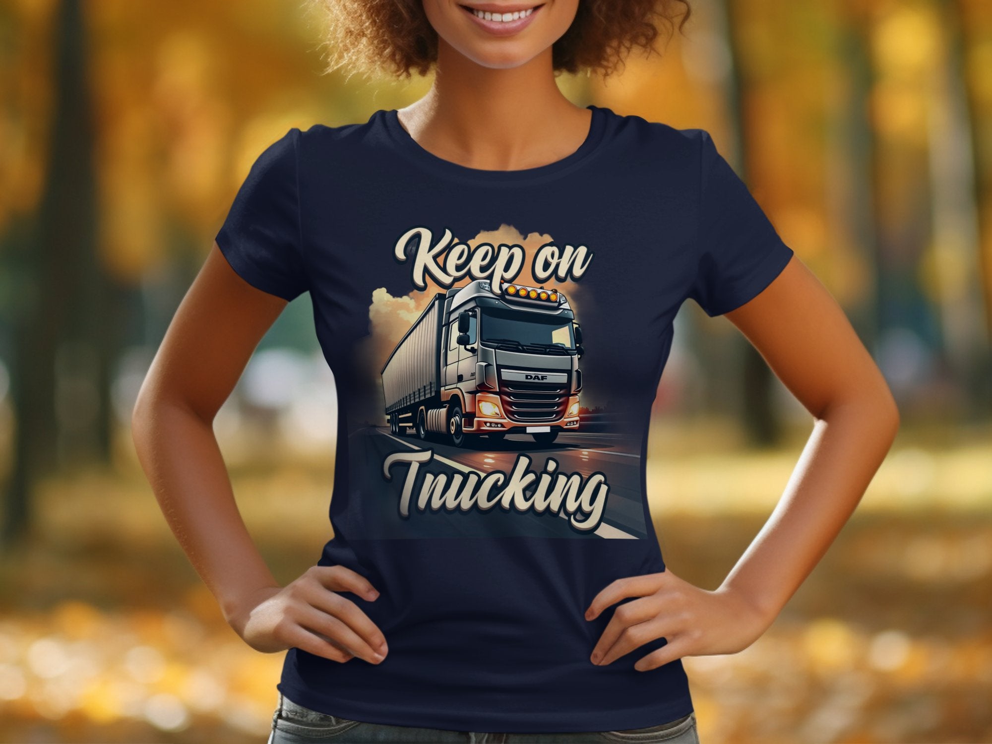 In a park, a person proudly dons a Garment Graphics navy blue T-shirt with a striking truck graphic and bold Keep on Trucking text, set against blurred warm-hued autumn foliage.