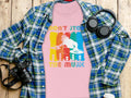 A flat lay image showcases a vibrant DJ graphic tee by Garment Graphics, featuring Dont Stop the Music text, ideal for music lovers. Its artfully arranged on a wooden surface with a blue and green plaid shirt, headphones, a camera, and a white bottle.