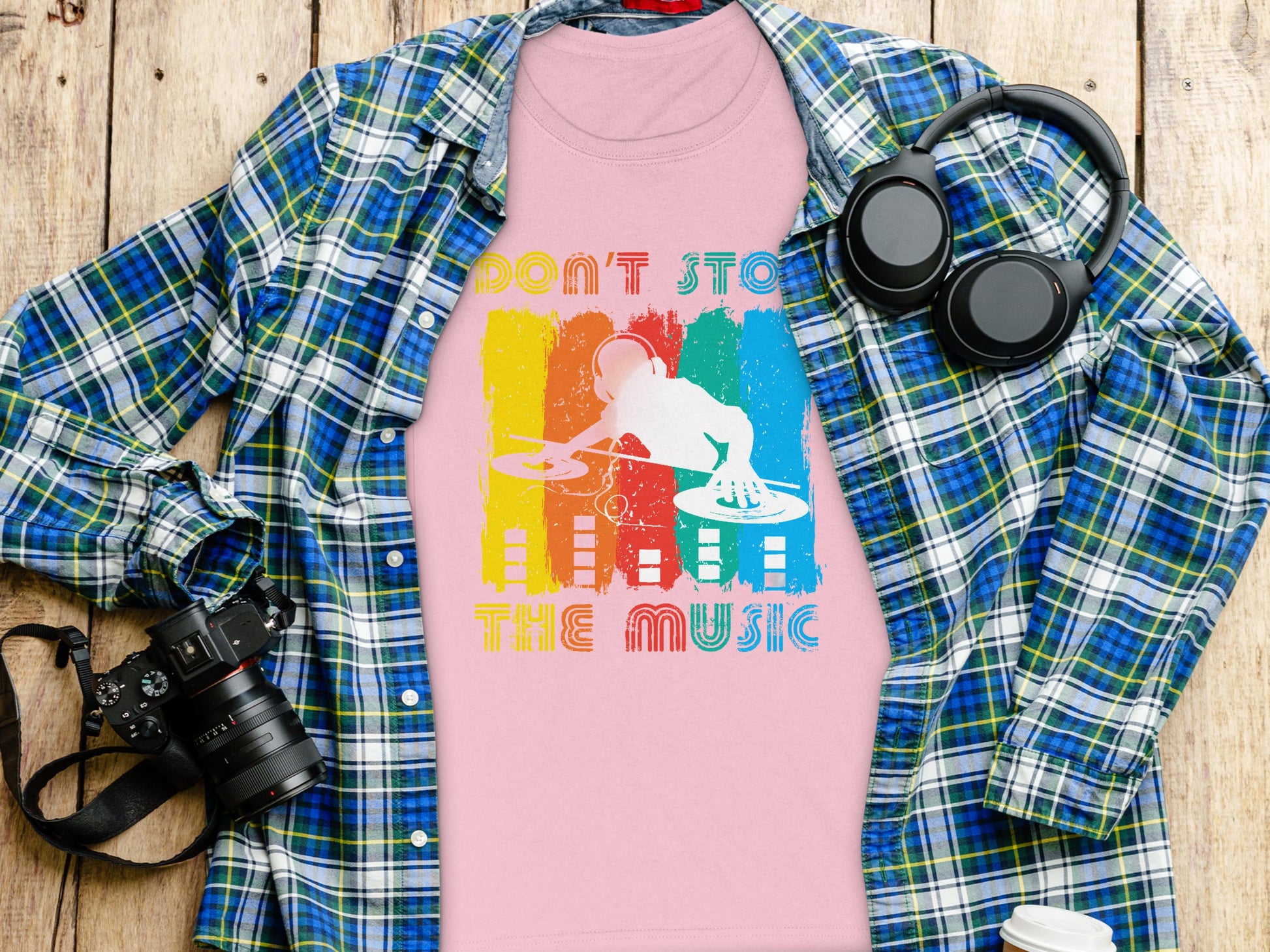 A flat lay image showcases a vibrant DJ graphic tee by Garment Graphics, featuring Dont Stop the Music text, ideal for music lovers. Its artfully arranged on a wooden surface with a blue and green plaid shirt, headphones, a camera, and a white bottle.