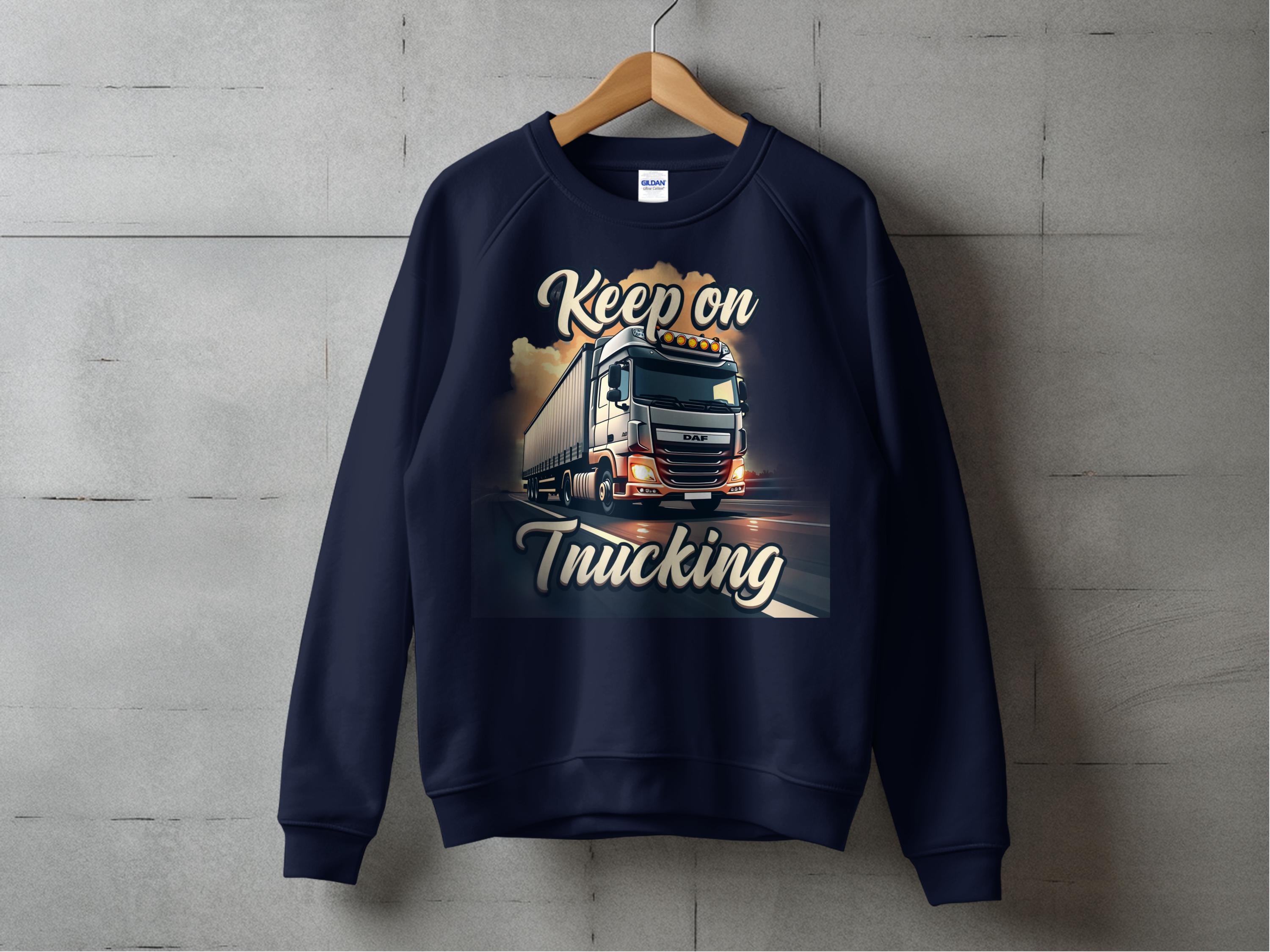 A navy blue Garment Graphics sweatshirt hangs on a wooden hanger against a concrete wall, featuring a vibrant large truck graphic with the message Keep on Trucking above and below, ideal for road enthusiasts seeking inspiration.