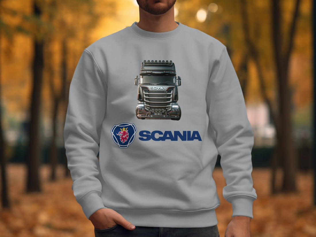 A Garment Graphics white T-shirt with a Scania truck design and logo lies on wood, alongside a blue plaid shirt, trendy sweatshirts, a camera, headphones, and a laptop—creating a casual and tech-savvy scene with graphic flair.
