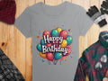 A gray, 100% cotton T-shirt by Garment Graphics features a vibrant Happy Birthday design with balloons, paired with a knitted hat, gloves, and plaid shirt all elegantly displayed on wood for a warm birthday celebration setup.
