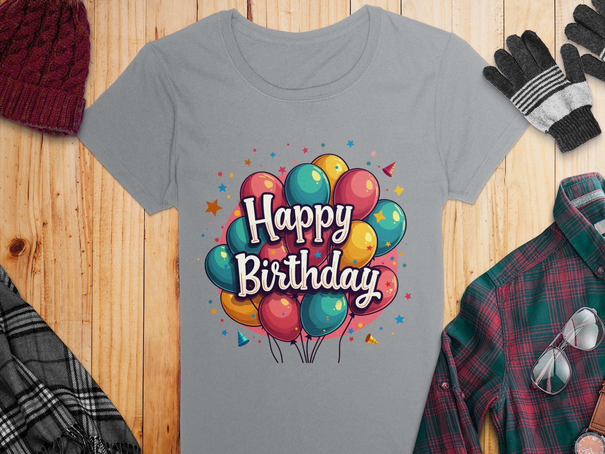 A gray, 100% cotton T-shirt by Garment Graphics features a vibrant Happy Birthday design with balloons, paired with a knitted hat, gloves, and plaid shirt all elegantly displayed on wood for a warm birthday celebration setup.