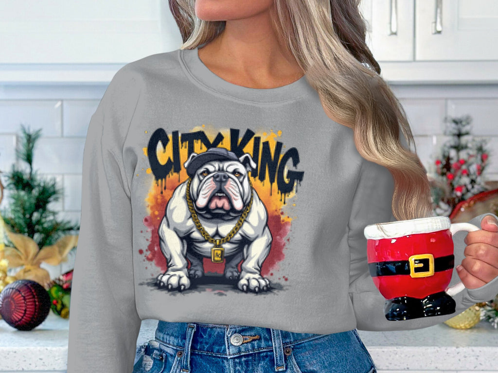 In a festive kitchen, someone holds a Santa-themed mug, wearing a Garment Graphics gray sweatshirt with a crowned bulldog and City King text. The surrounding holiday greenery and ornaments perfectly complement their stylish appearance.