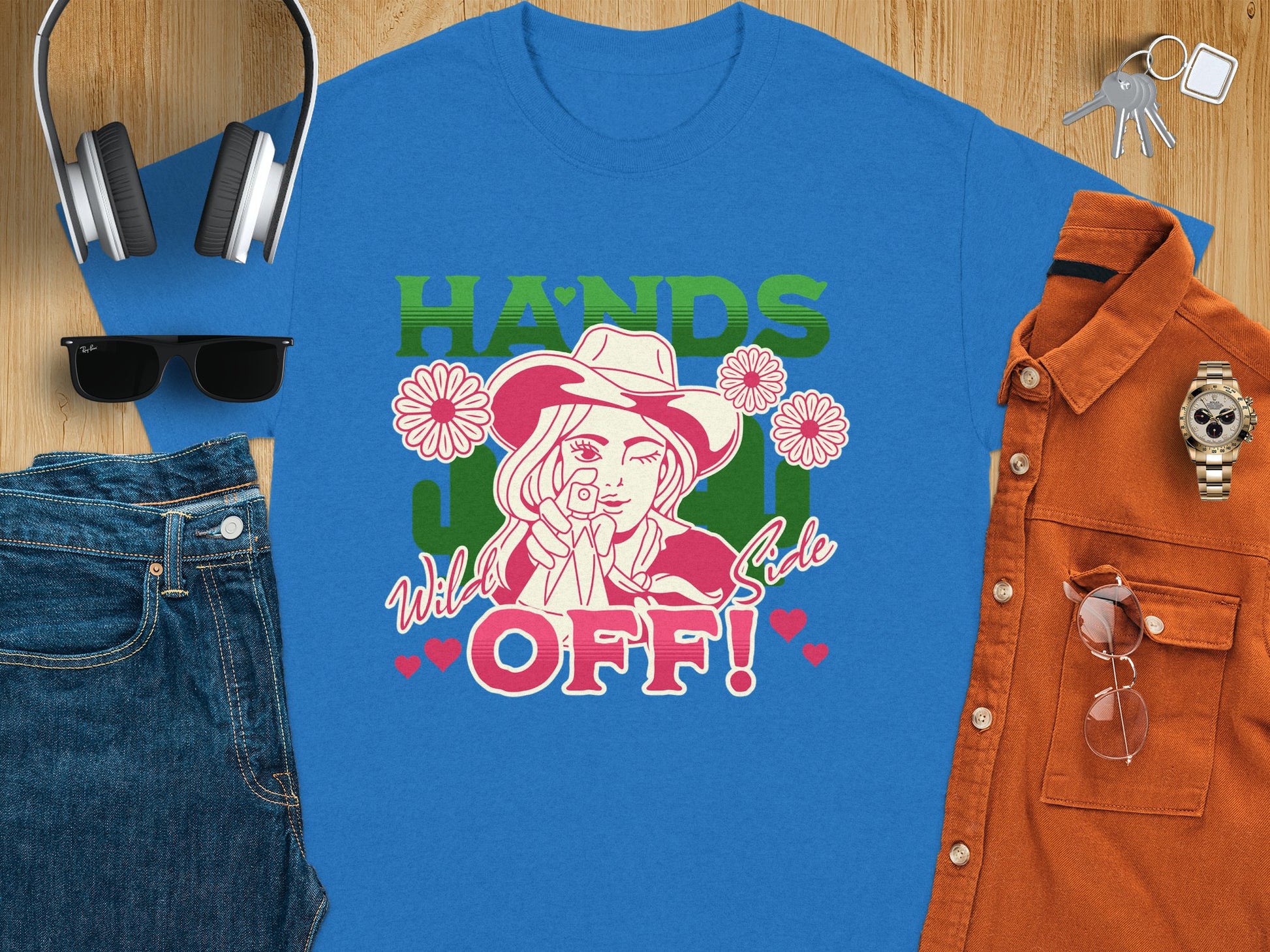 Garment Graphics presents a blue t-shirt showcasing a striking design of a woman in a cowboy hat and bandana. HANDS OFF! is adorned with flowers and hearts. This classic fit pairs seamlessly with sunglasses, headphones, denim, a rust shirt, keys, and a watch.