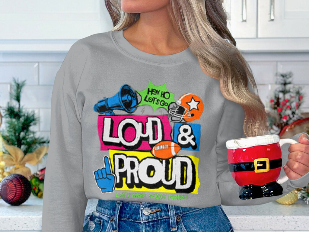 In a kitchen adorned with holiday decor, someone holds a Santa-themed mug, wearing a Garment Graphics gray Loud & Proud Sweatshirts top embellished with vibrant megaphone, football, and foam finger graphics—showcasing their distinct style.