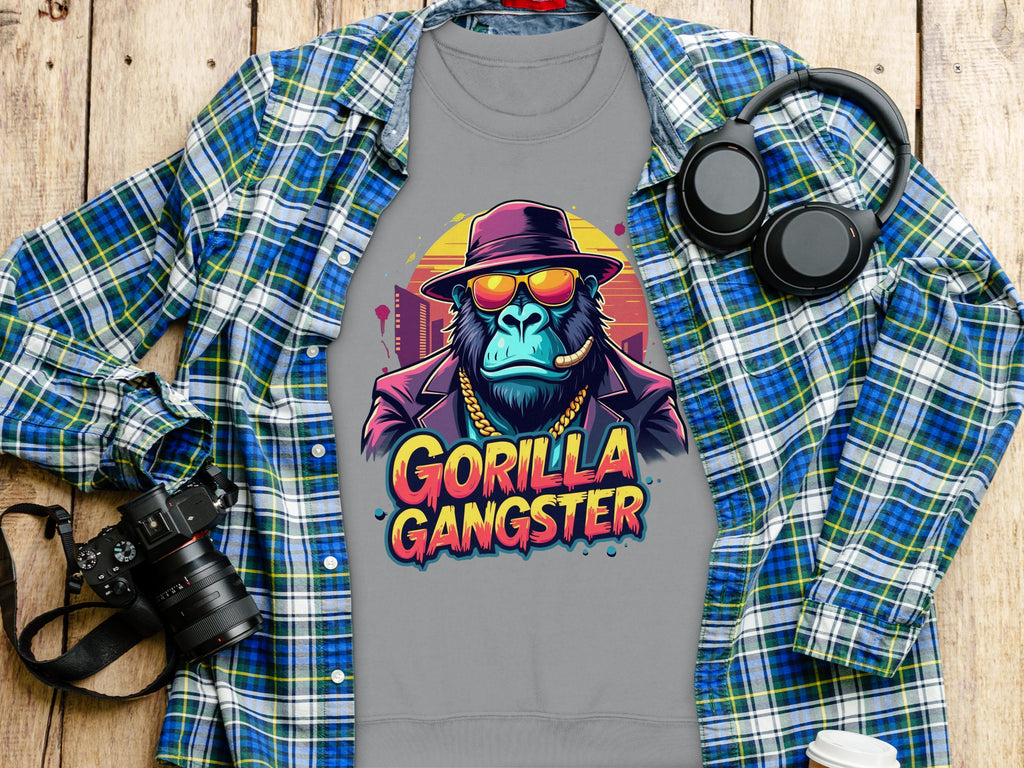A gray t-shirt from Garment Graphics features a Urban Style Gorilla Gangster design with a cartoon gorilla in sunglasses and a hat, placed on a wooden surface next to a blue plaid shirt, black headphones, camera, and white cup.