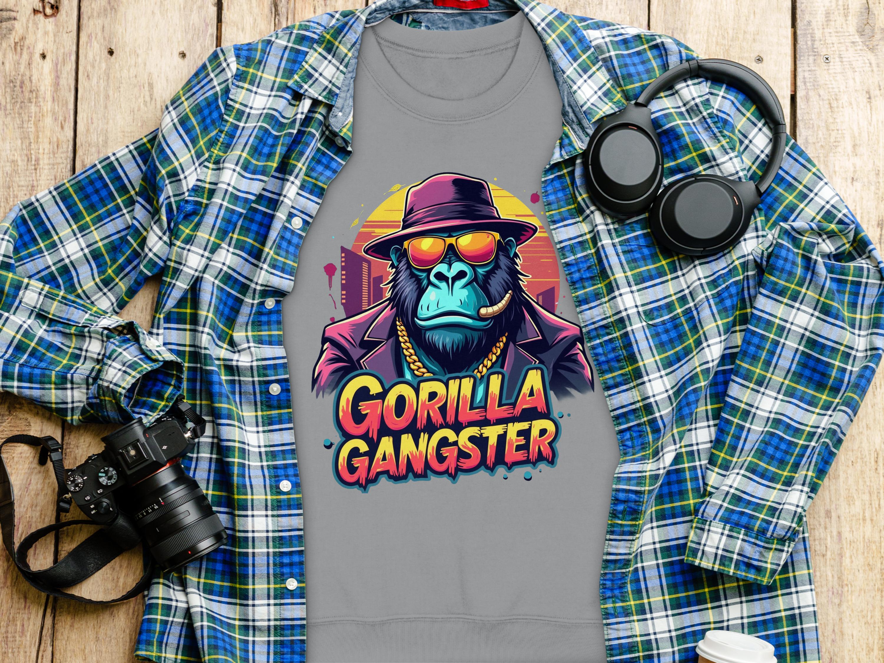 A gray t-shirt from Garment Graphics features a Urban Style Gorilla Gangster design with a cartoon gorilla in sunglasses and a hat, placed on a wooden surface next to a blue plaid shirt, black headphones, camera, and white cup.