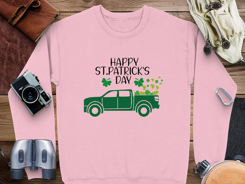 A Garment Graphics pink sweatshirt features Happy St. Patricks Day above a green truck filled with shamrocks, styled on a wooden surface with accessories like a watch, camera, binoculars, belt, and leather item for an adventurous festive display.