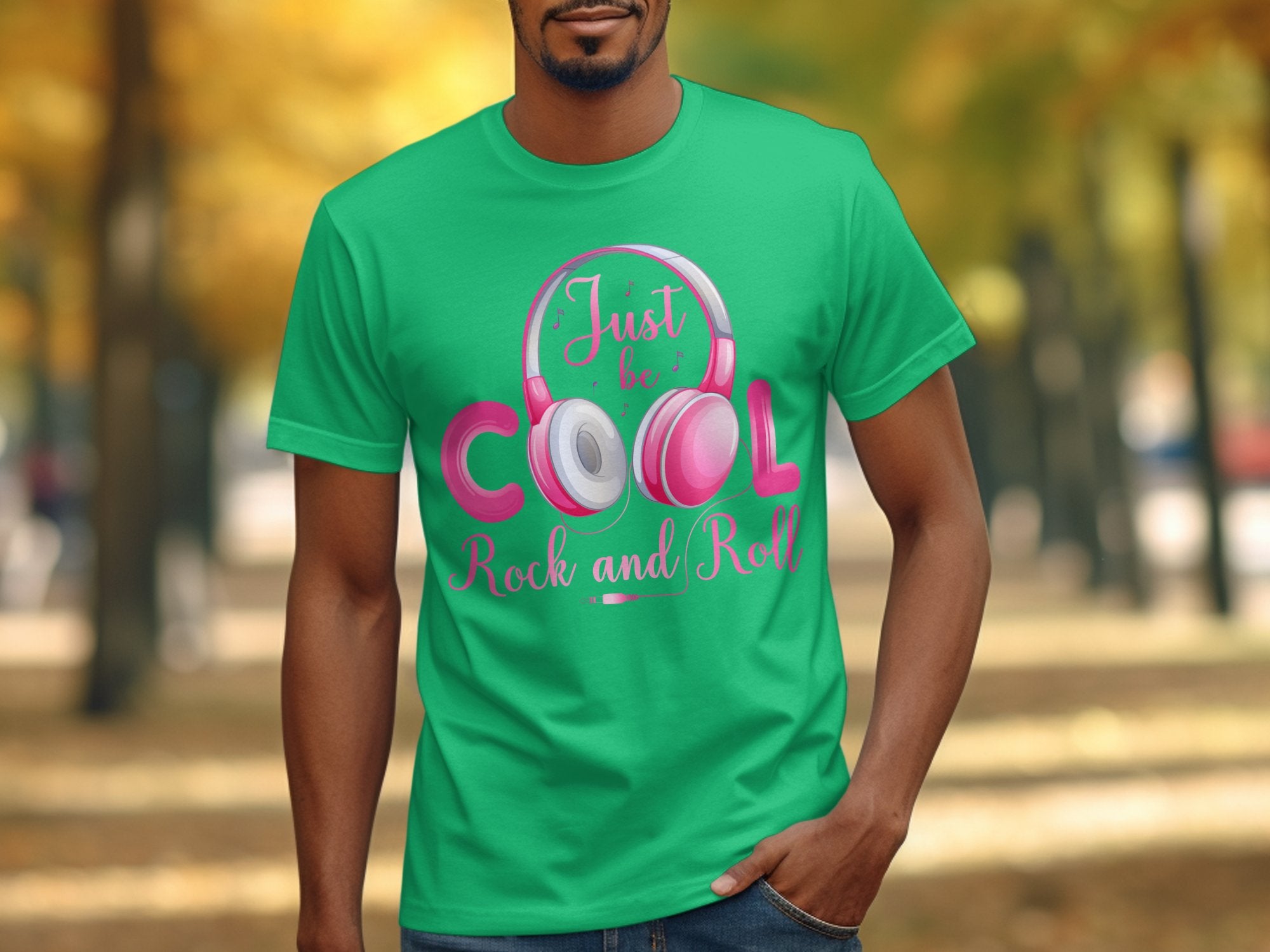 A person outdoors in a green T-shirt by Garment Graphics featuring headphones and Just be cool, rock and roll text is surrounded by blurry autumn trees. Ideal for music lovers who enjoy trendy Rock and Roll designs.