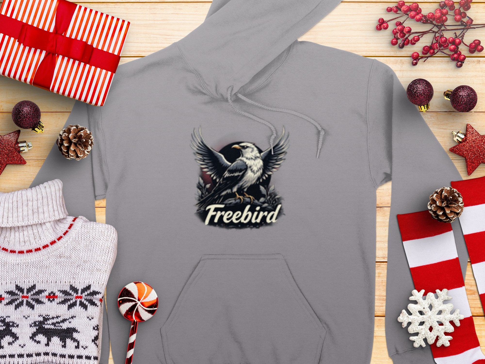 Celebrate the festive season in style with the Garment Graphics Freebird Graphic Pullover Hoodie, featuring an eagle and Freebird text. Surrounded by red and white wrapped gifts, pine cones, a knitted sweater, and peppermint candies, its unique bird design enhances your holiday vibe.