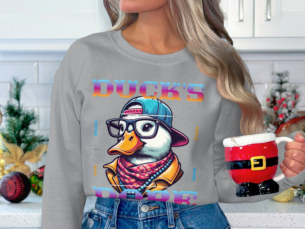A person holds a Santa-themed mug while wearing Garment Graphics stylish sweatshirt. It features a colorful duck illustration with glasses and a cap, titled Ducks Dope. Kitchen cabinets in the background are adorned with festive holiday decorations, making a bold fashion statement.