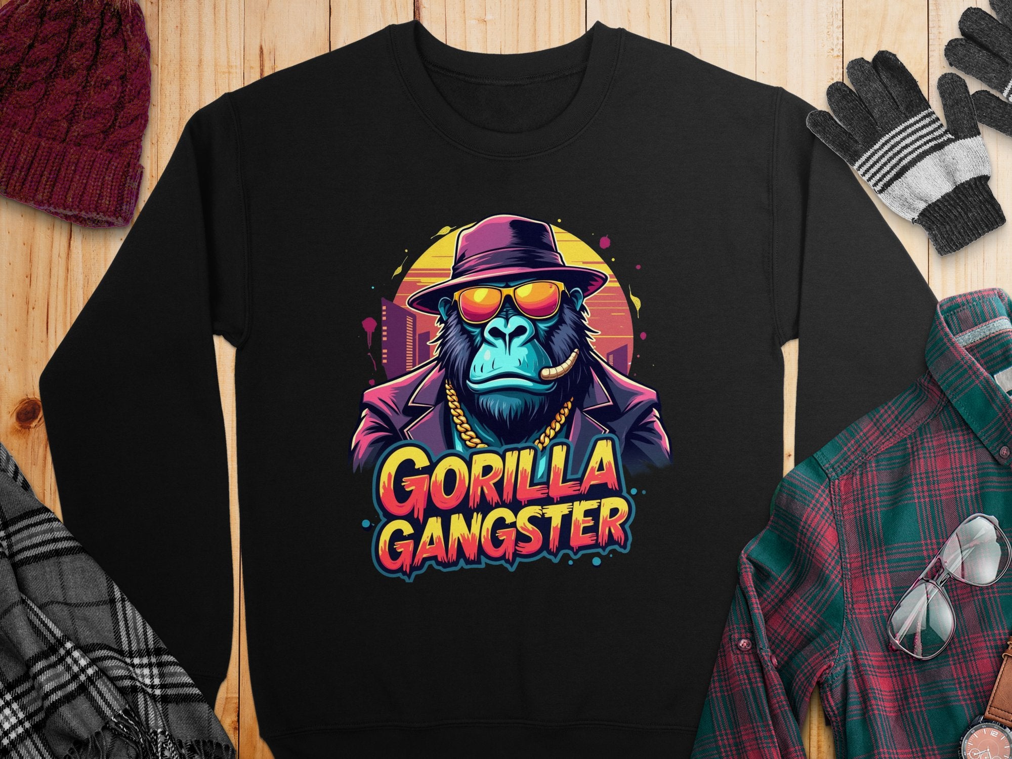 Garment Graphics urban-style sweatshirt features a gorilla with a hat, sunglasses, and necklace labeled Gorilla Gangster. It complements gloves, a scarf, and flannel shirt seamlessly for the ultimate street-savvy look against a wooden surface.