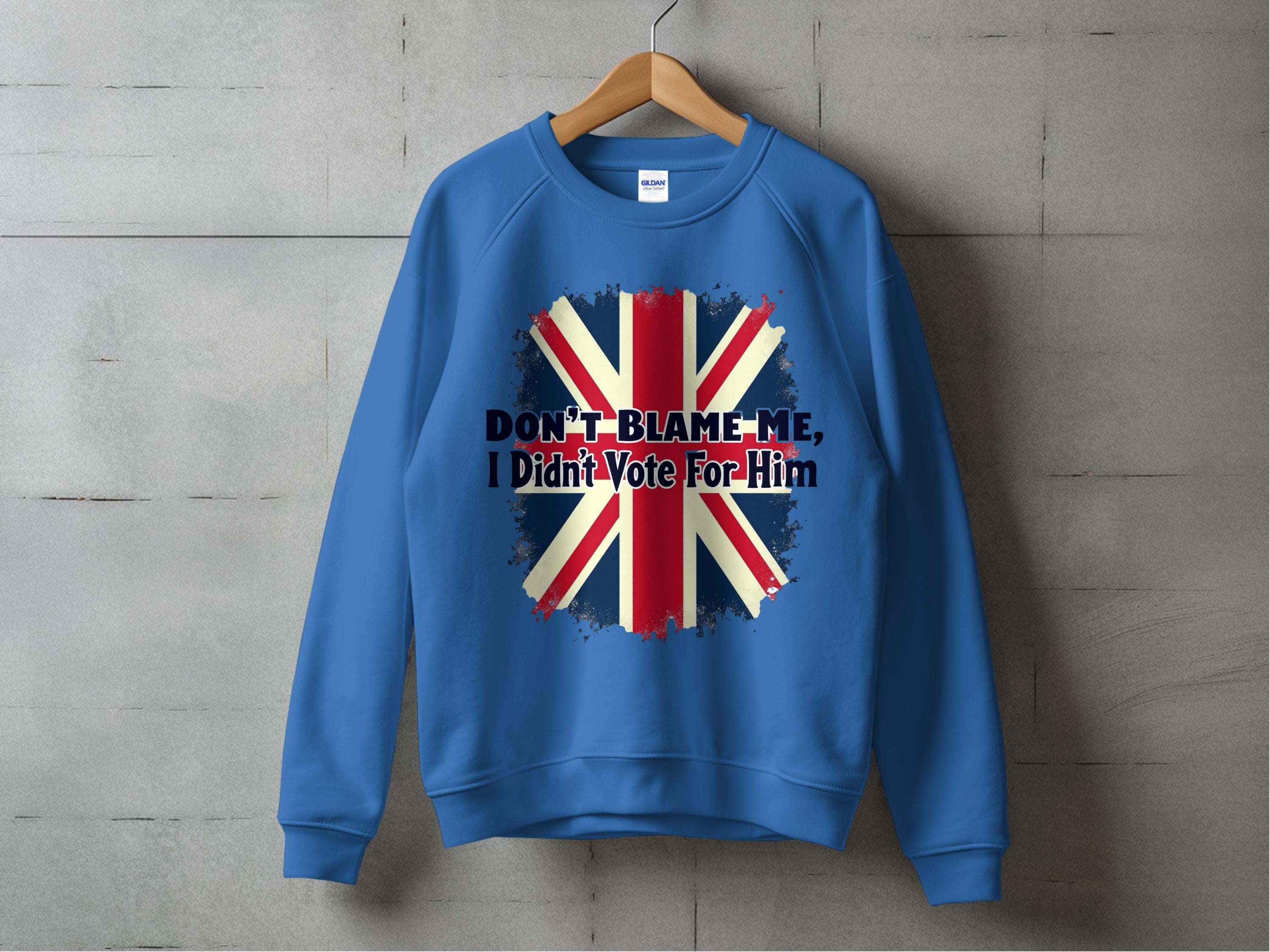 A blue unisex sweatshirt by Garment Graphics hangs on a wooden hanger against a concrete wall. It showcases a distressed Union Jack and the bold statement Dont Blame Me, I Didnt Vote For Him, making it a striking addition to any wardrobe.