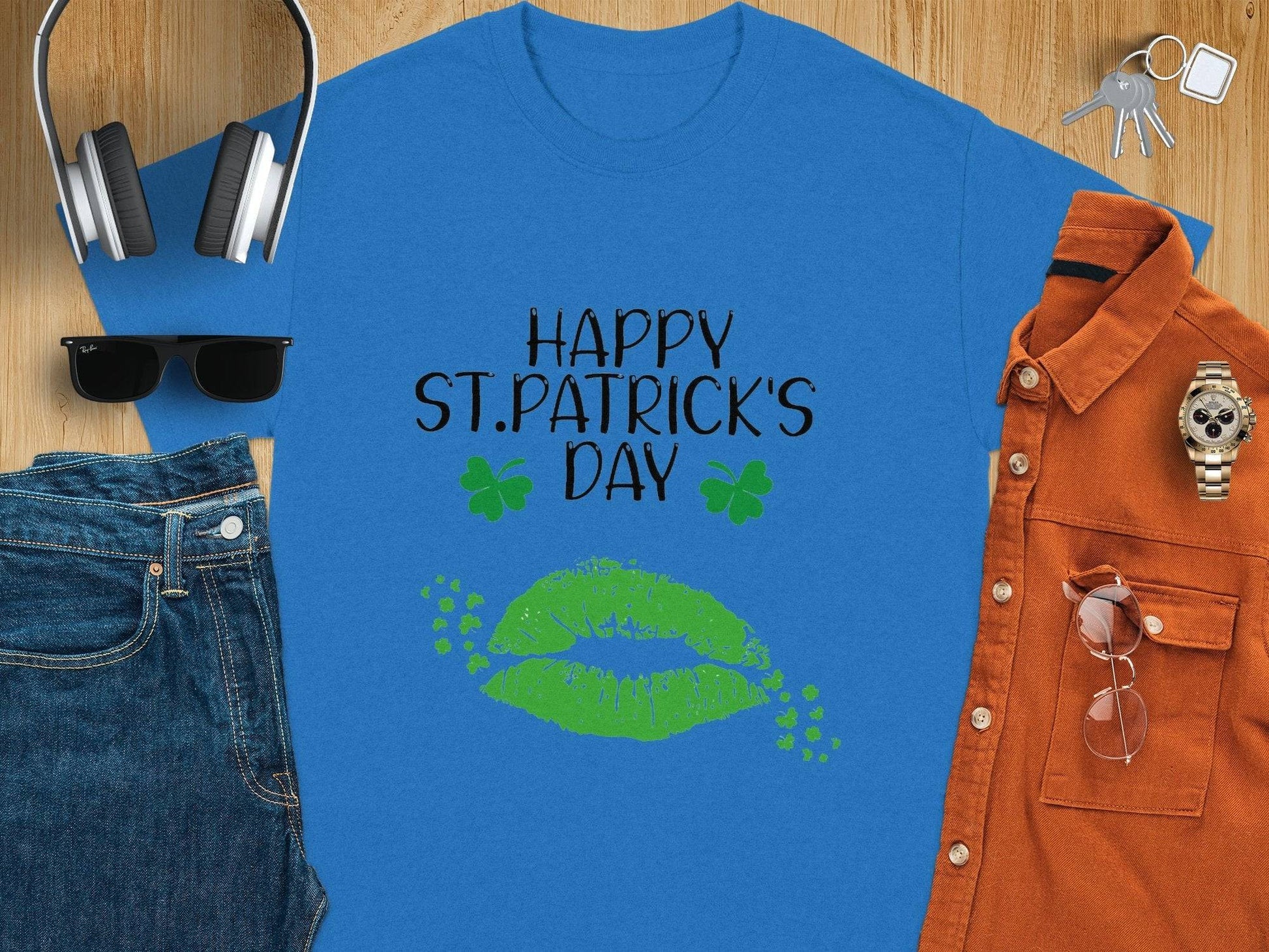 The Happy St. Patricks Day graphic T-shirt by Garment Graphics is blue with black text, two green shamrocks, and vibrant green lips. Its styled with sunglasses, denim jeans, a watch, an orange shirt, headphones, a cap, and keys on wood.