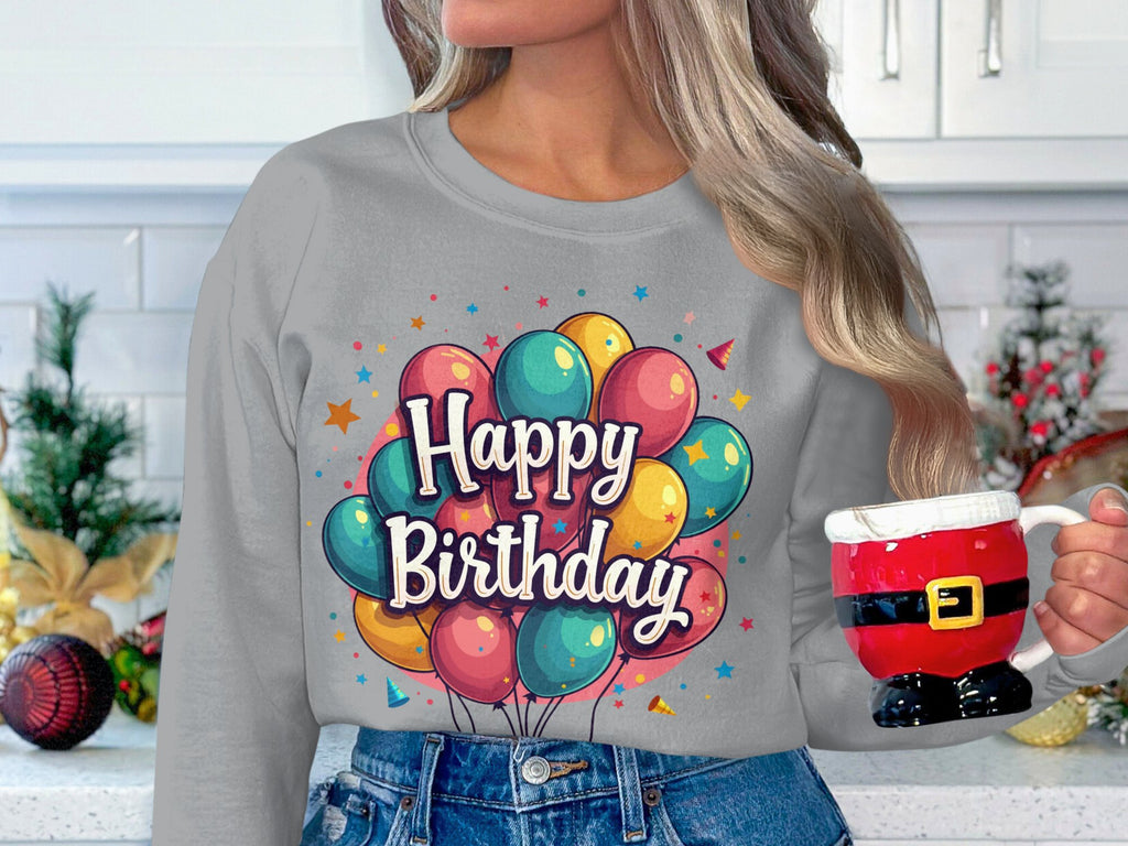 In a festive kitchen, someone wears a Garment Graphics gray Happy Birthday sweatshirt, featuring colorful balloons and unisex sizing. Theyre holding a mug styled like Santas suit, with holiday decorations adorning the counter in the background.
