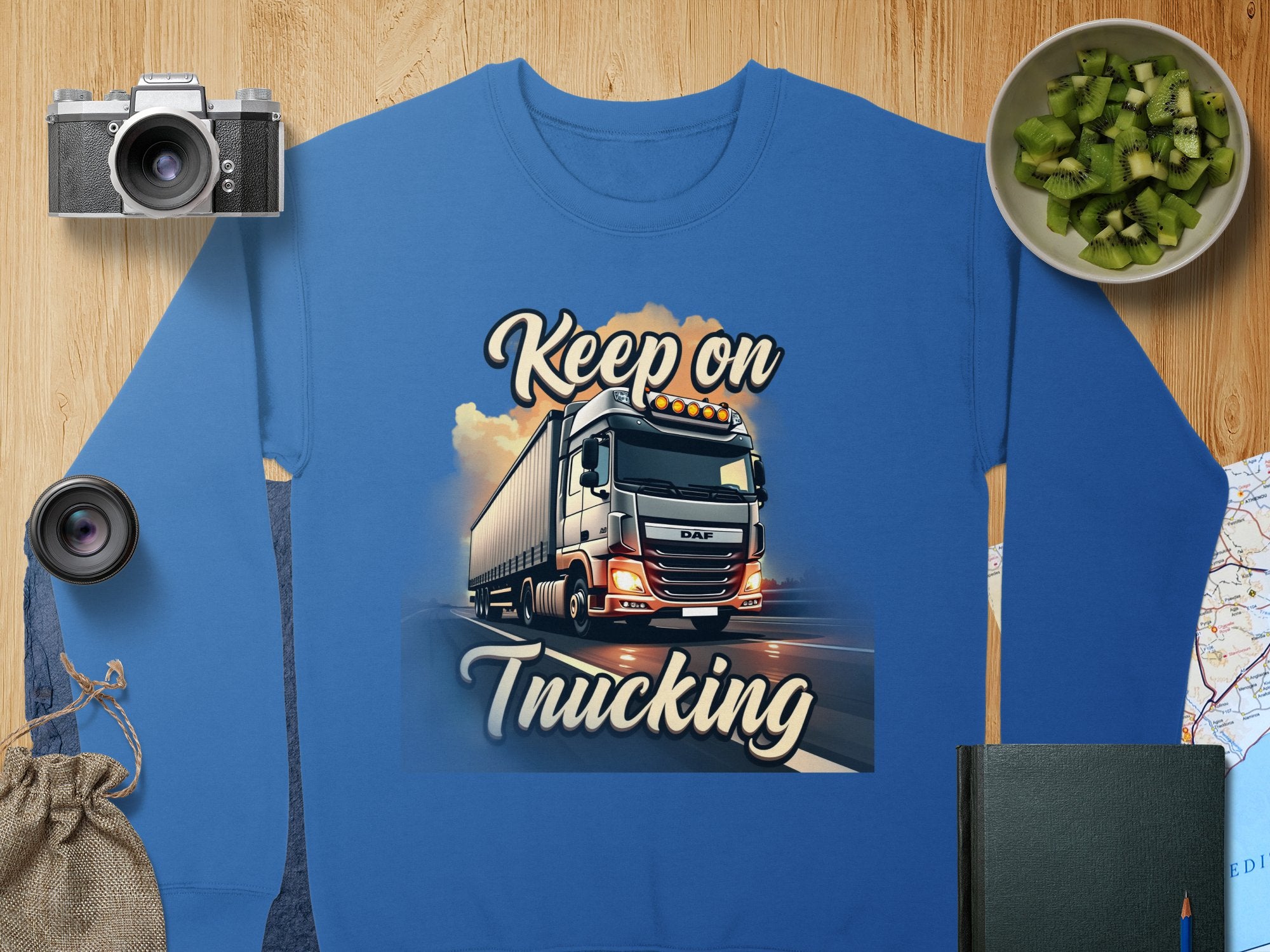 Garment Graphics presents a unisex blue graphic sweatshirt with a large truck and Keep on Trucking text. Displayed on wood with a camera, lens, sliced kiwi, book, pouch, and map for an adventure-ready vibe.