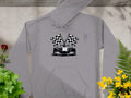 Garment Graphics gray hoodie showcases a graphic race car with a checkered flag design. Ideal for motorsport fans, this stylish piece is displayed on a gray surface surrounded by potted plants with red and yellow flowers.