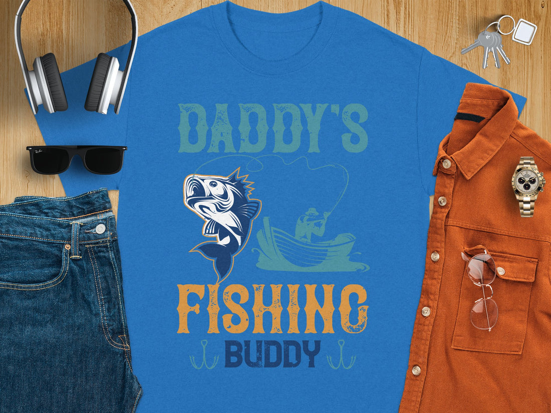 A person wears a Garment Graphics t-shirt, featuring Daddys Fishing Buddy with a fun fish and boater scene, decorative hooks, and set against a sandy background with hints of grass. Perfect for fishing fans.