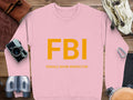 A Garment Graphics pink sweatshirt with bold yellow FBI and smaller Female Boob Inspector text is displayed on a wooden surface, surrounded by a camera, binoculars, a leather strap, and a canteen. This fun unisex piece is perfect for everyone.