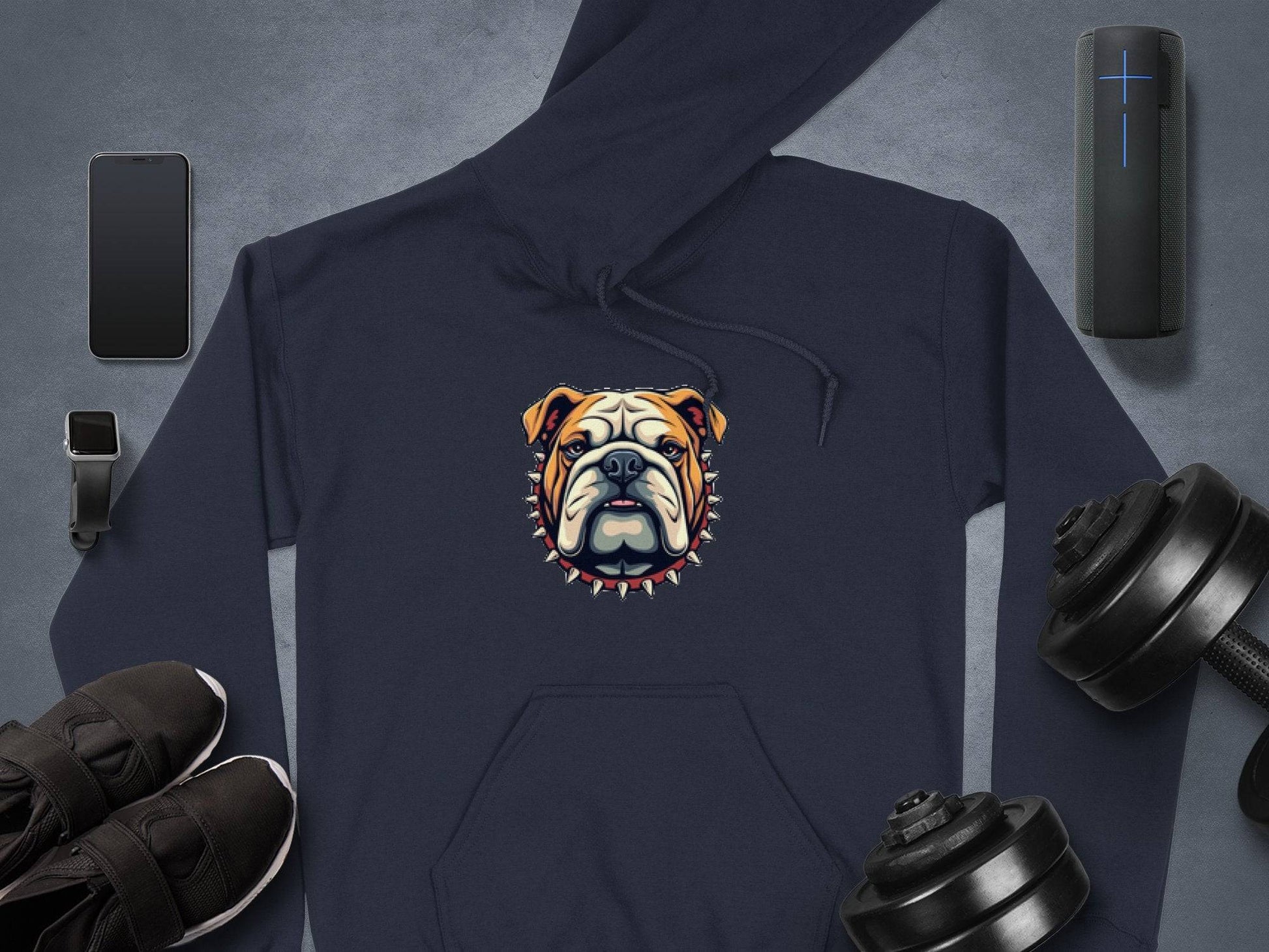 A navy hoodie featuring a bold bulldog graphic with a spiked collar from Garment Graphics lies on a gray surface, surrounded by a smartphone, smartwatch with band, wireless speaker, black sneakers, and two dumbbells.