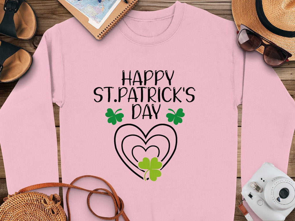 Garment Graphics showcases a pink festive sweatshirt with Happy St. Patricks Day in black lettering, adorned with green shamrocks and clover designs. Nearby are shoes, a notebook, a hat, sunglasses, and a camera.