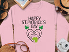 Garment Graphics showcases a pink festive sweatshirt with Happy St. Patricks Day in black lettering, adorned with green shamrocks and clover designs. Nearby are shoes, a notebook, a hat, sunglasses, and a camera.