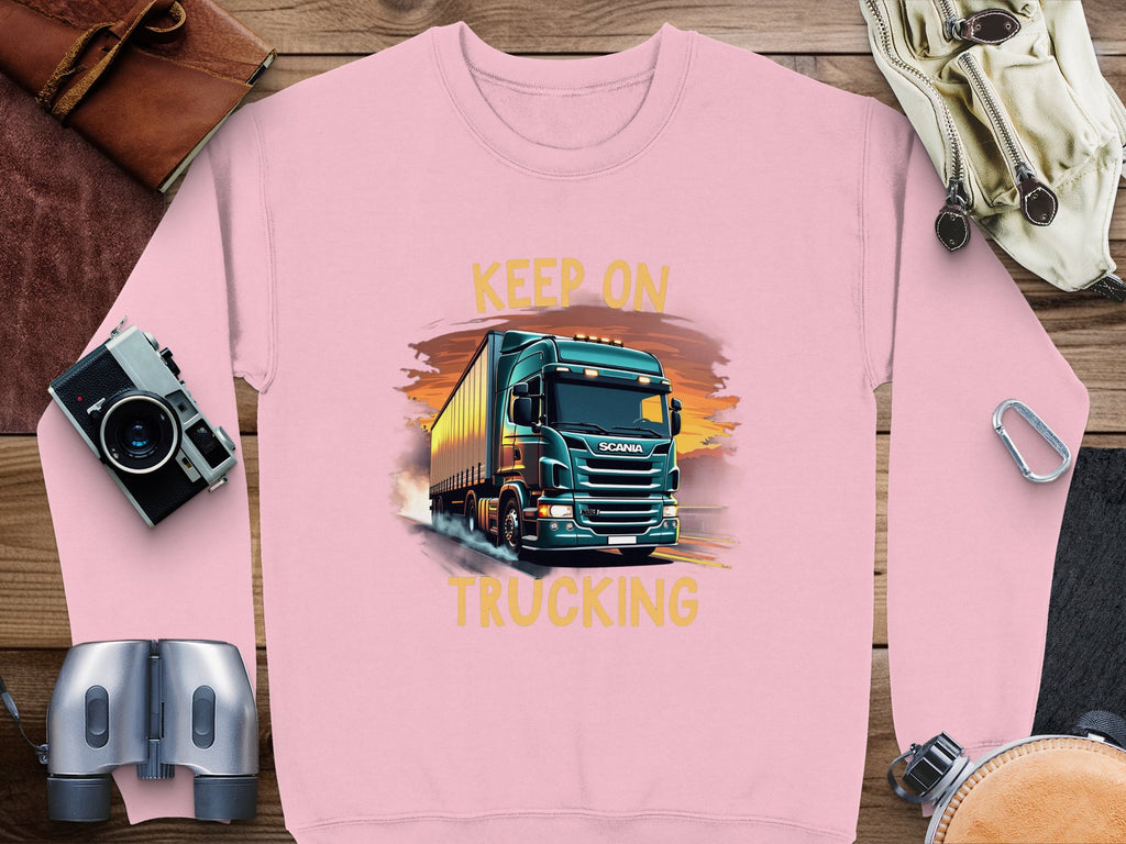 A pink Garment Graphics sweatshirt features a large truck illustration with KEEP ON TRUCKING above it, ideal for truck lovers. Nearby are a camera, binoculars, diary, sunglasses, and headphones on wood.