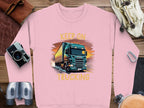 A pink Garment Graphics sweatshirt features a large truck illustration with KEEP ON TRUCKING above it, ideal for truck lovers. Nearby are a camera, binoculars, diary, sunglasses, and headphones on wood.