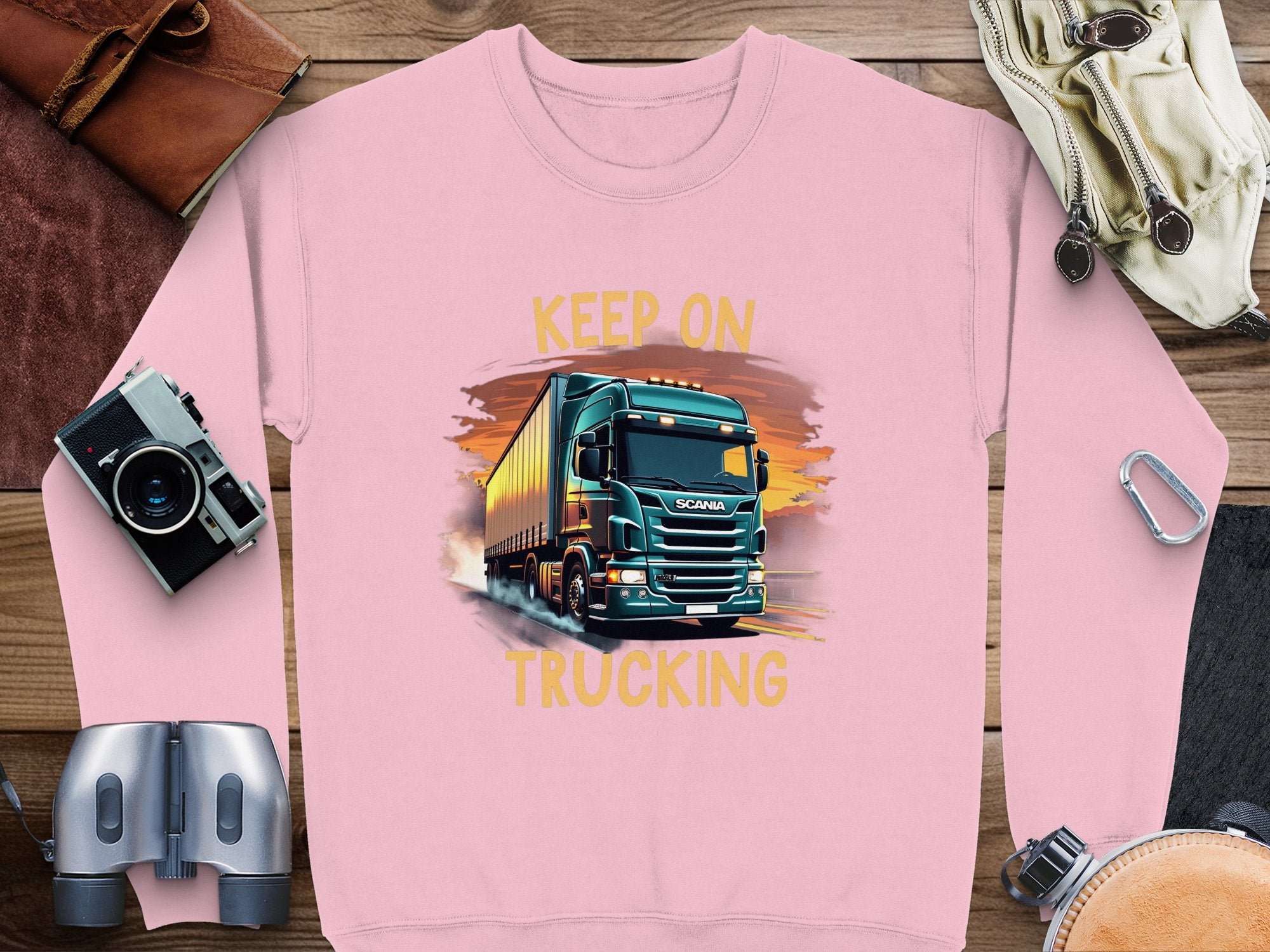 A pink Garment Graphics sweatshirt features a large truck illustration with KEEP ON TRUCKING above it, ideal for truck lovers. Nearby are a camera, binoculars, diary, sunglasses, and headphones on wood.
