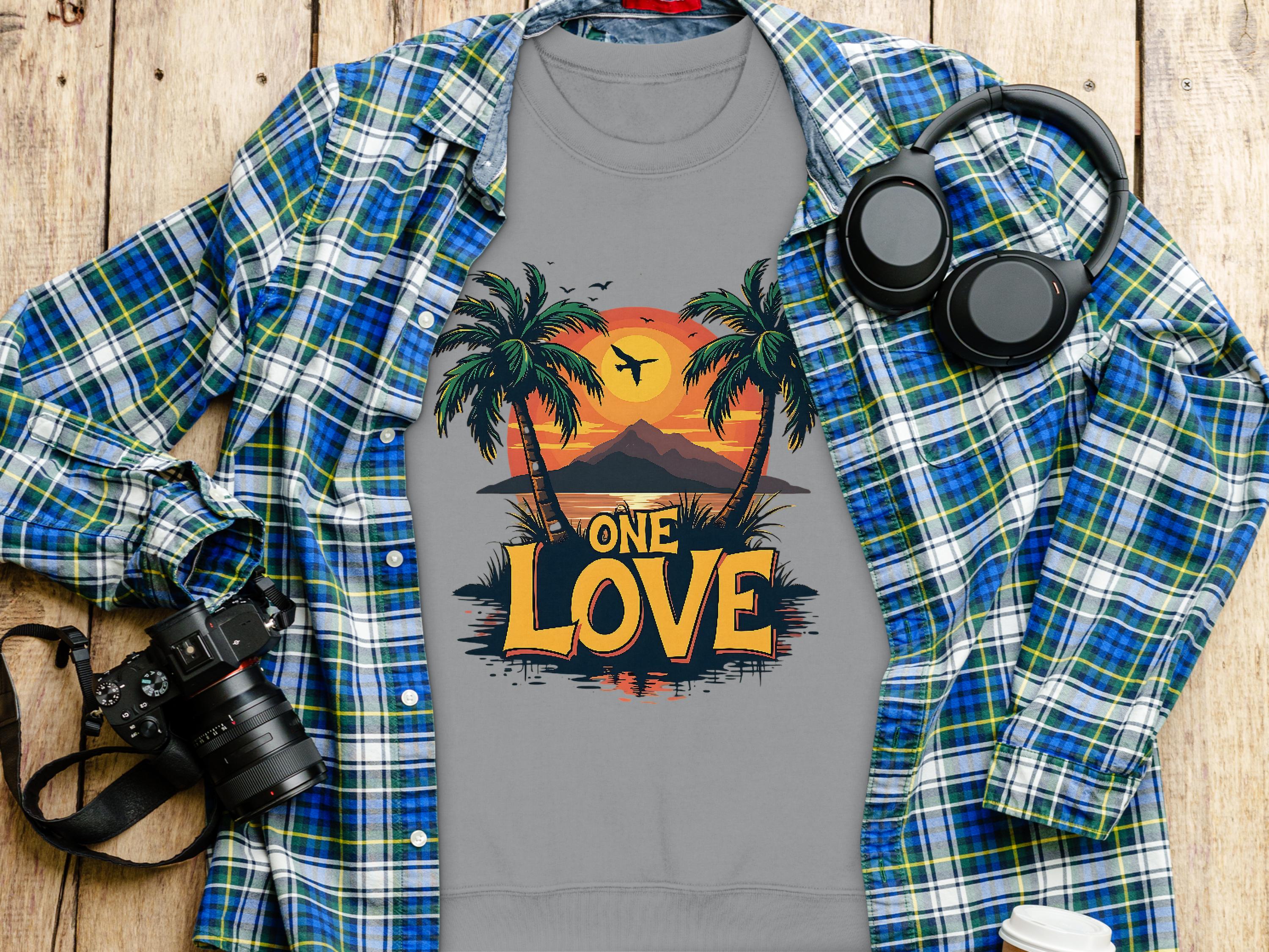 A Garment Graphics plaid blue and green shirt over a gray One Love Sunset T-shirt with palm trees. A camera, headphones, pen, and coffee cup lid sit artfully arranged on the wooden surface.