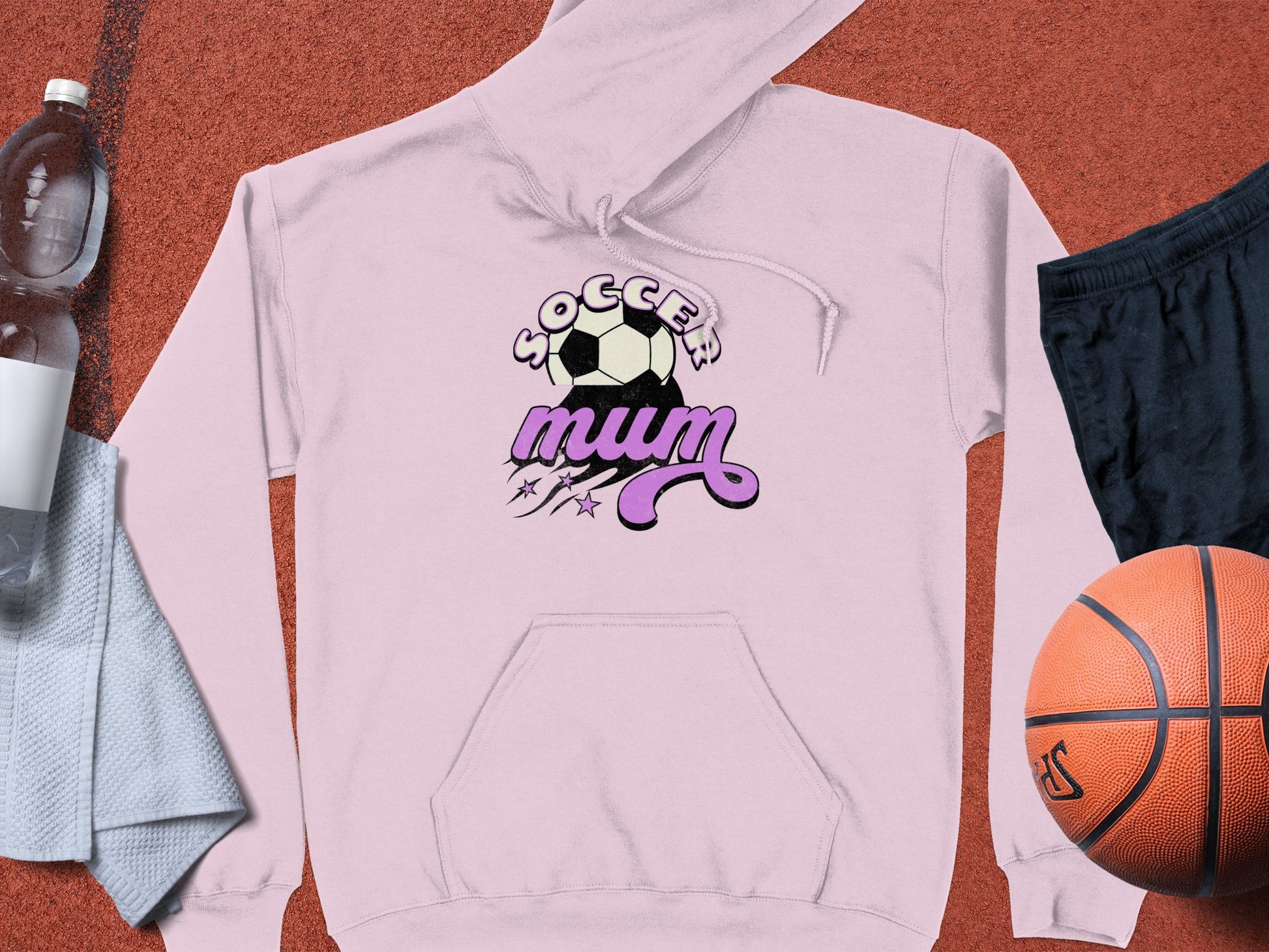 Garment Graphics presents a cozy pink Soccer Mum hoodie with a soccer ball design. Nearby, a basketball, black shorts, water bottle, and towel lie on a vibrant red surface—perfect for sports enthusiasts.