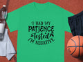 A green T-shirt from Garment Graphics with I had my patience tested Im negative in black stylized text rests on a textured brown surface, surrounded by a water bottle, gray towel, black shorts, and an orange basketball.