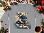 A gray Garment Graphics sweatshirt from the Graphic Inspirational Collection features a Keep on Trucking truck, surrounded by Christmas decor like pine branches and ornaments with a red star, all set on wood next to a steaming coffee cup.