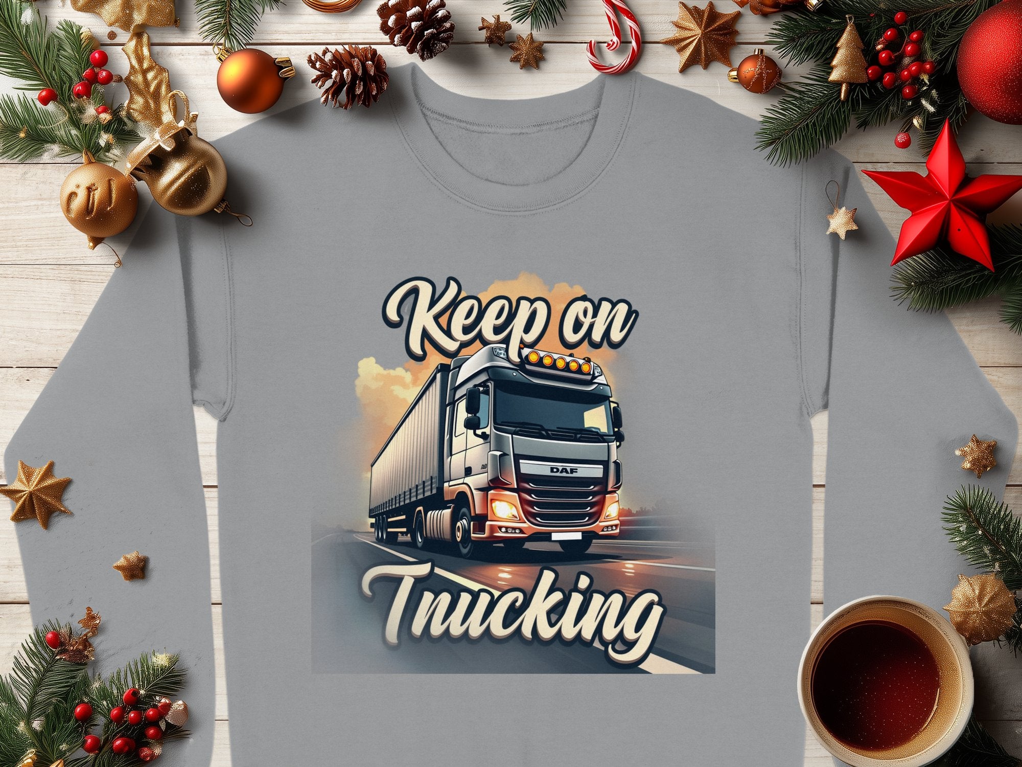 A gray Garment Graphics sweatshirt from the Graphic Inspirational Collection features a Keep on Trucking truck, surrounded by Christmas decor like pine branches and ornaments with a red star, all set on wood next to a steaming coffee cup.