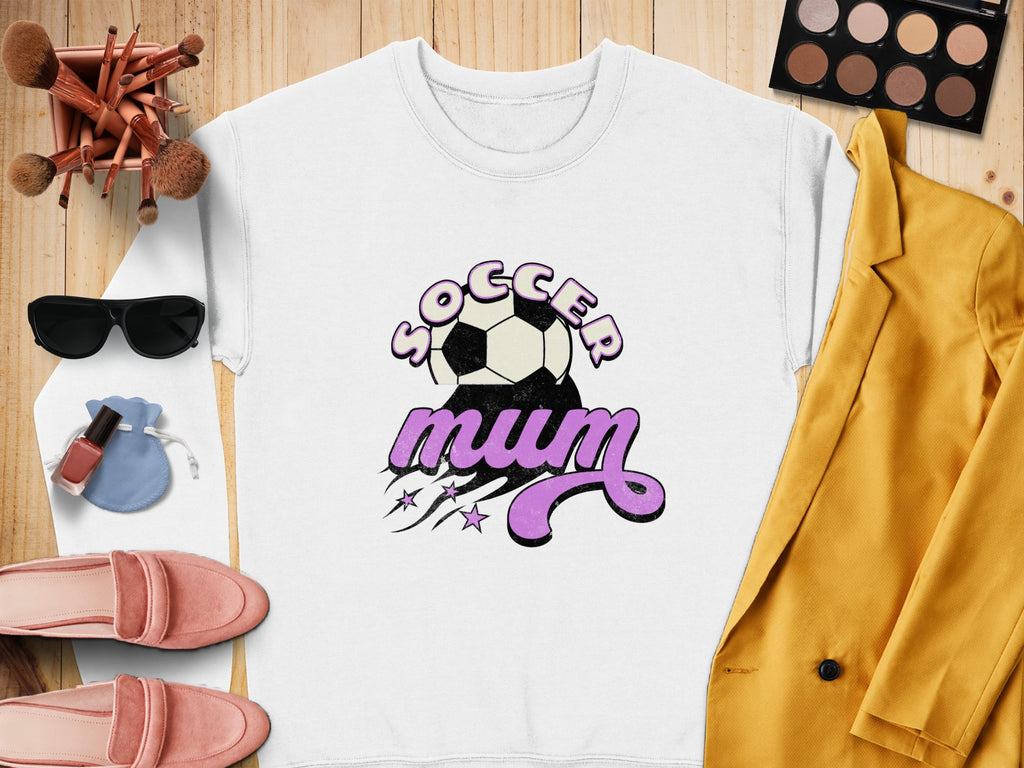 This vintage-style white sweatshirt from Garment Graphics displays Soccer Mum in bold purple text alongside a soccer ball graphic. It’s stylishly placed on wood, accompanied by chic makeup, sunglasses, pink shoes, a yellow blazer, and a vibrant blue pouch.