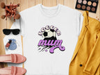 This vintage-style white sweatshirt from Garment Graphics displays Soccer Mum in bold purple text alongside a soccer ball graphic. It’s stylishly placed on wood, accompanied by chic makeup, sunglasses, pink shoes, a yellow blazer, and a vibrant blue pouch.