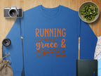 A blue Garment Graphics sweatshirt with Running on grace & coffee in bold orange text is spread out on a wooden surface, surrounded by a camera, lens, chopped kiwi bowl, drawstring bag, notebook, and part of a map. This versatile piece offers unisex sizing for everyone.