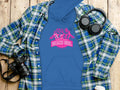 Flat lay featuring a Garment Graphics blue Always Party Beach Girl hoodie with vibrant pink text, paired with a blue and green plaid shirt. Black headphones, a black camera, and a white cup complete the casual style on the wooden surface.