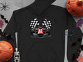 A Garment Graphics black hoodie features a graphic of a red racing car with checkered flags. Its surrounded by two pumpkins, black spiders, bats, a small skeleton figurine, and autumn leaves on a gray surface.