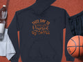 Garment Graphics navy coffee hoodie with This Day is Powered by Coffee in cursive rests stylishly on a basketball court. Its paired with black shorts, an orange basketball, a white towel, and a clear plastic water bottle—ideal for active coffee lovers.