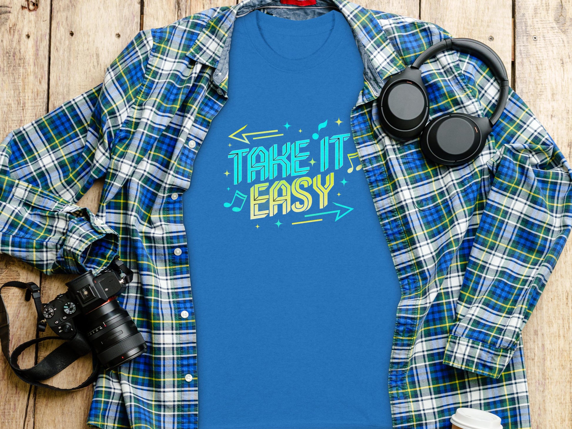 A vibrant blue Take It Easy T-shirt with playful musical notes and arrows by Garment Graphics lies on a wooden surface, surrounded by a green and blue plaid shirt, a camera, headphones, and a coffee cup.