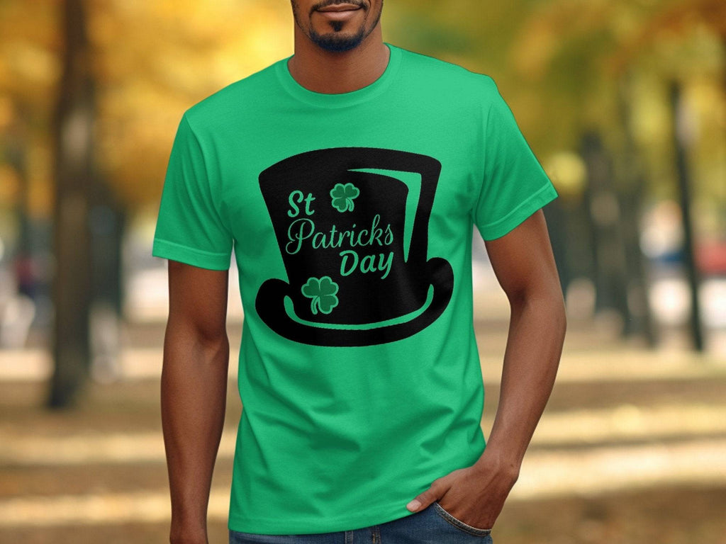In an outdoor autumn park, a person wears a bright green Garment Graphics T-shirt with a black top hat graphic, St Patricks Day text, and two shamrocks, contrasting the colorful trees. The festive design includes a four-leaf clover.
