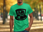 In an outdoor autumn park, a person wears a bright green Garment Graphics T-shirt with a black top hat graphic, St Patricks Day text, and two shamrocks, contrasting the colorful trees. The festive design includes a four-leaf clover.