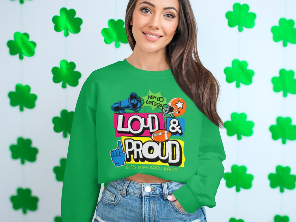 A woman stands out in a green Garment Graphics sweatshirt, featuring Hey Ho Lets Go Loud & Proud Lets Make Some Noise text with lively designs. The unisex piece is part of the Loud and Proud Sweatshirts collection. Shamrock decorations adorn the white wall behind her.