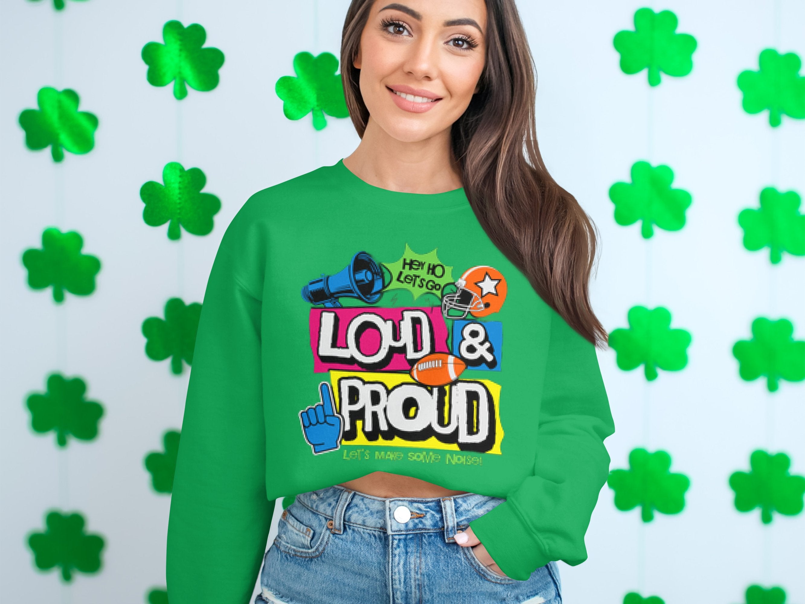 A woman stands out in a green Garment Graphics sweatshirt, featuring Hey Ho Lets Go Loud & Proud Lets Make Some Noise text with lively designs. The unisex piece is part of the Loud and Proud Sweatshirts collection. Shamrock decorations adorn the white wall behind her.