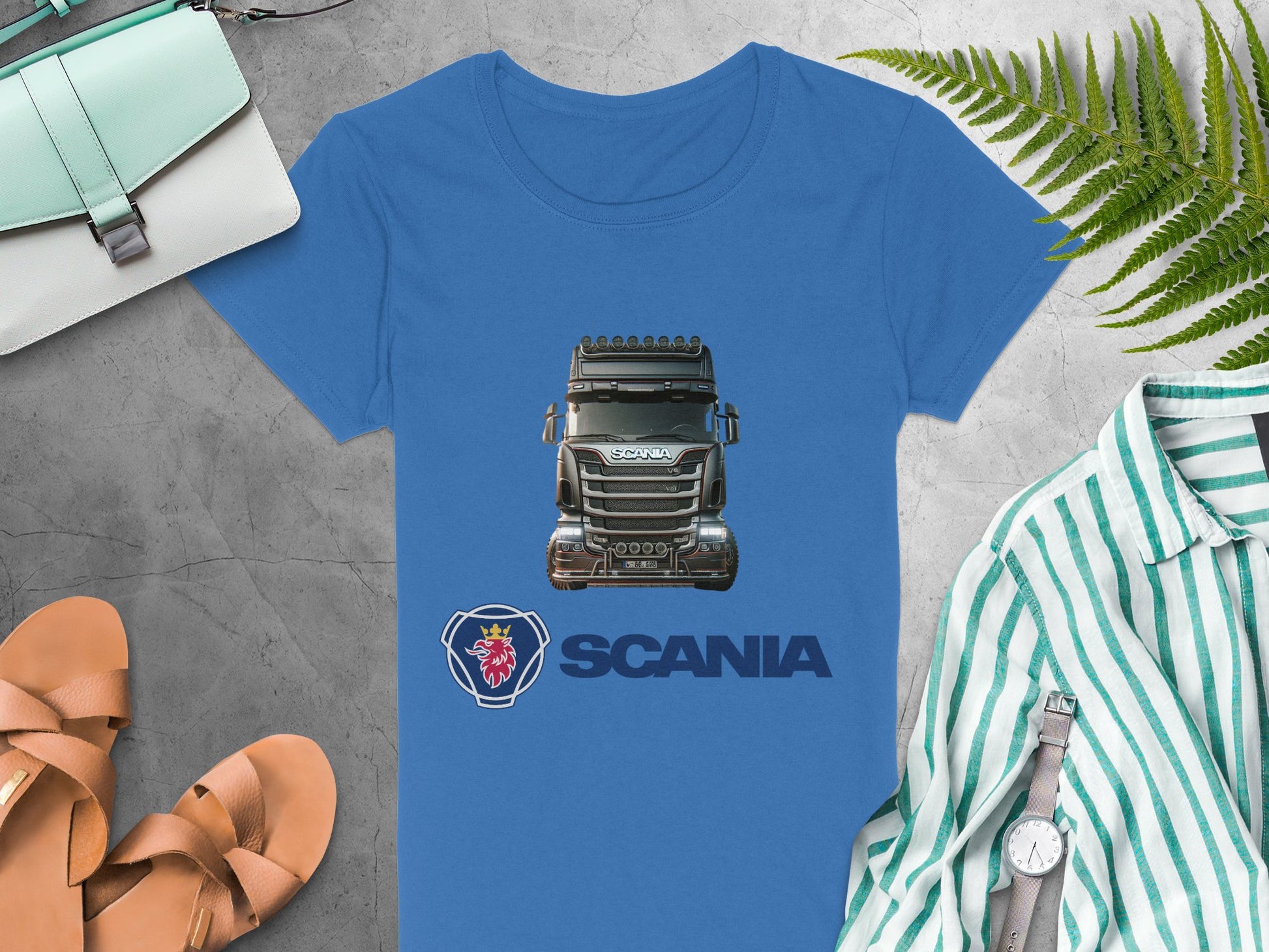 Garment Graphics blue Scania T-shirt, featuring a truck image and logo, is displayed on a gray surface. Ideal for automotive enthusiasts, its accompanied by sandals, a turquoise bag, striped shirt, wristwatch, and fern leaf.