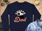 Garment Graphics navy sweatshirt features Soccer Dad with Soccer above a retro soccer ball and Dad below in cursive, laid flat on wood with a denim jacket draped nearby.